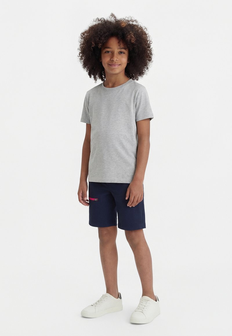 Παιδί με σγουρά μαλλιά στέκεται χαμογελαστό, φοράει γκρι κοντομάνικο μπλουζάκι, ναυτικά σορτς με τσέπη με φερμουάρ και άσπρα αθλητικά παπούτσια μπροστά σε λευκό φόντο.