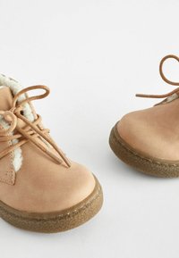 Bottes en cuir tan à lacets avec une doublure douce de couleur crème, semelles en caoutchouc texturé et orteils arrondis. Détails de couture visibles.