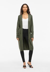Long cardigan vert olive avec deux poches avant, ouvert sur le devant et avec des poignets côtelés, porté par-dessus un haut blanc et un pantalon skinny noir.