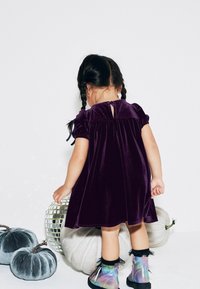 Vestido de terciopelo púrpura con mangas abullonadas y detalle de ojal en la espalda. Combinado con botas de tobillo iridiscentes y rodeado de calabazas decorativas.