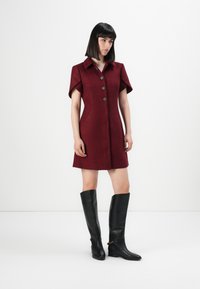 Donna in un vestito bordeaux a maniche corte con bottoni, abbinato a stivali al ginocchio in pelle nera con dettagli in catena dorata.