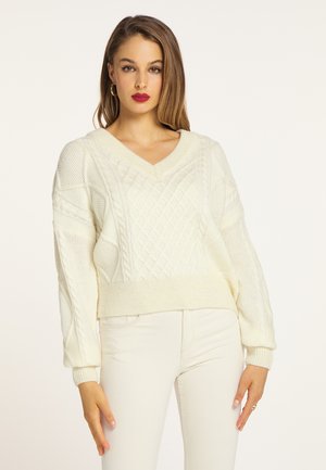 Vrouw met lang bruin haar, gekleed in een crèmekleurige kabelgebreide V-hals trui en witte broek, staand tegen een lichte achtergrond.