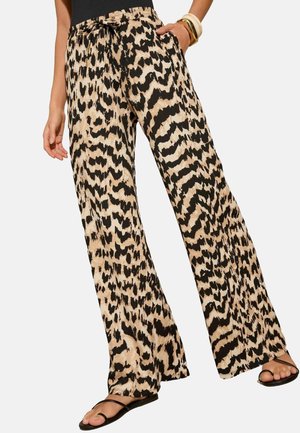 Femme portant un pantalon large à imprimé animal beige et noir avec ceinture à nouer, assorti de sandales noires et de bracelets au poignet.