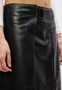 Μαύρη δερμάτινη φούστα με φαρδύ λάστιχο στη μέση, λεπτομέρεια ζώνης και αντίθεση ραφής. Διαθέτει εφαρμοστή γραμμή και απαλό υφή.