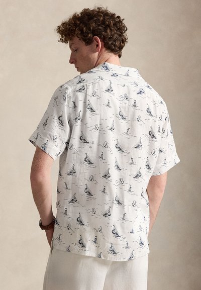 Homme portant une chemise blanche à manches courtes avec des motifs de voiliers et d’ancres bleu marine, tourné pour montrer le dos de la chemise.