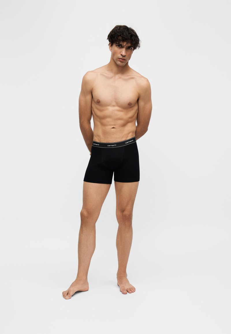 Junger Mann mit lockigem Haar steht barfuß in schwarzen Carhartt Boxershorts vor einem schlichten hellen Hintergrund.