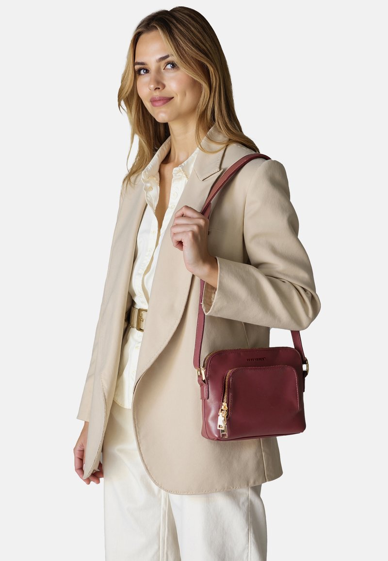 Borsa a tracolla in pelle bordeaux con tasca con zip, dettagli in metallo dorato e tracolla regolabile, tenuta da una persona con un blazer beige.