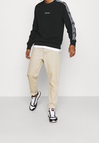 Felpa nera con logo bianco, pantaloni beige a sigaretta e sneakers bianco e nere con dettagli rossi. Presenta un orlo e polsini a costine.