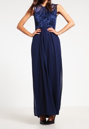 Mujer de pie con las manos en las caderas, vestida con un vestido largo azul marino sin mangas con corpiño de encaje y falda fluida.