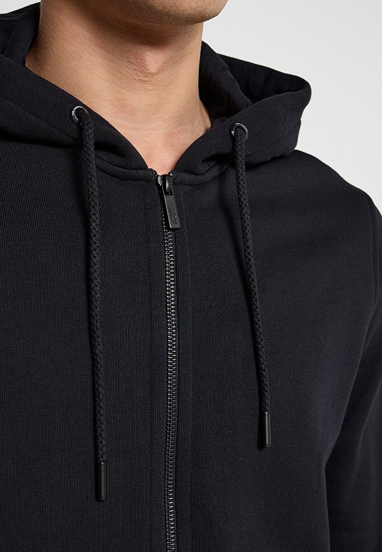 Felpa nera con zip realizzata in tessuto a costine. Presenta un cappuccio con lacci e una cerniera metallica con accenti in tessuto. Vista ravvicinata del colletto.