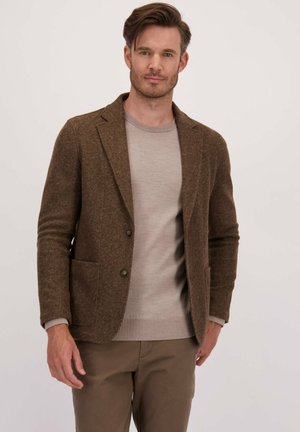 Man in een bruine tekstuur blazer, lichtbeige sweater en bruine broek, staande tegen een effen lichte achtergrond.