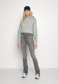 Suéter cinza de malha com gola alta, combinado com jeans cinza ajustados e ténis brancos. O modelo também usa um boné preto.