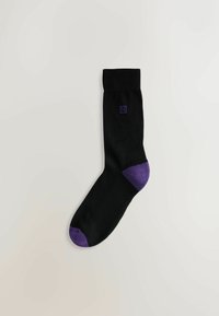 Schwarze Socke mit violetten Akzenten an Zehen und Ferse. Hergestellt aus Stoff mit glatter Textur. Mit einem kleinen lila Logo versehen.
