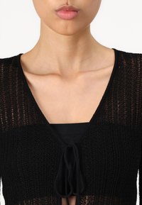 Cardigan nero lavorato a maglia con scollatura a V, caratterizzato da una vestibilità slim, tessuto testurizzato e chiusura anteriore con fiocco. La maglietta sottostante è nera.
