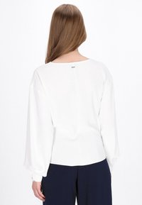 Blusa bianca a maniche lunghe con scollatura rotonda, texture liscia e una sottile cucitura sul retro, abbinata a pantaloni blu scuro. Accento in hardware argentato.