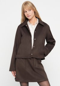 Bruine, getextureerde stoffen jas met een brede kraag en drukknoopsluitingen, gecombineerd met een bijpassende rok. Glad, eenvoudig ontwerp, zonder patronen.