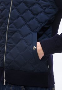 Μπλε ναυτικό μπουφάν με καπιτονέ υφή, ασημένια τσέπη με κούμπωμα, πλευρικό φερμουάρ και ριμπ μανσέτες, φορεμένο με το χέρι μέσα στην τσέπη και σκούρο τζιν.