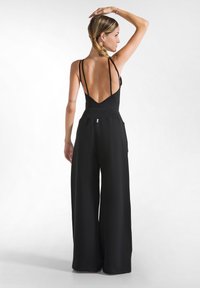 Mono negro con un escote en V profundo en la espalda, tirantes finos y un diseño de pierna ancha. Hecho de una tela suave, con bolsillos y un detalle mínimo.