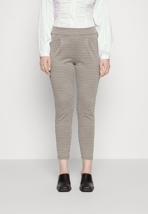 Femme debout portant un pantalon ajusté beige et noir à carreaux, un chemisier blanc plissé et des chaussures noires sans lacets, sur un fond uni.