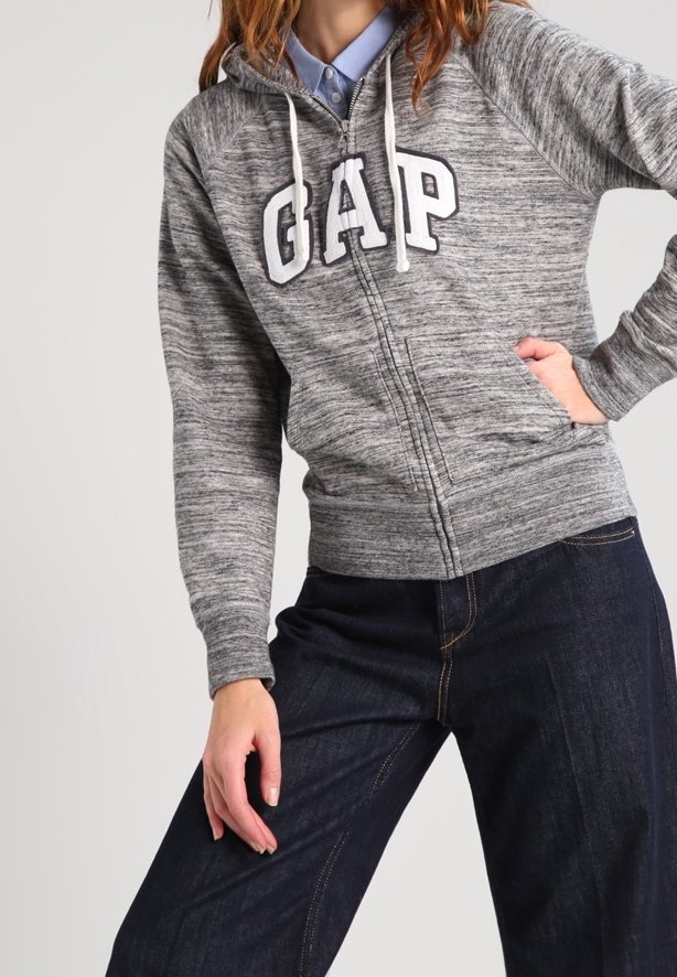 Felpa grigia con zip e texture in pile, con un grande logo bianco "GAP" sul petto e stringhe sul cappuccio. Indossata con jeans scuri.
