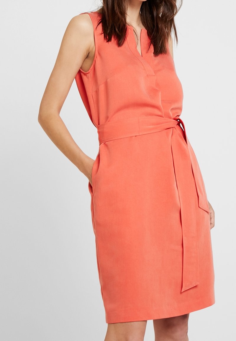 Robe corail sans manches avec un col en V, une taille ceinturée et des poches latérales, confectionnée dans un tissu lisse à texture mate.