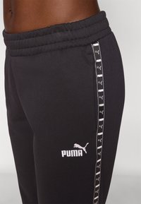 Schwarze Jogginghose mit einem elastischen Bund, featuring ein weißes Puma-Logo und einen vertikalen gepunkteten Streifen an der Seite. Weiche Stofftextur.