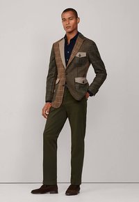 Geruite blazer in bruin- en groene stof met contrasterende tankleurige stukken, gecombineerd met olijfgroene broek en bruine schoenen. Knopdetails op de zakken.