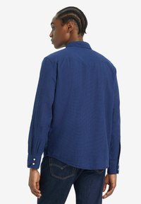 Camisa de cuadros azul marino con mangas largas, detalles de botones en los puños y un cuello clásico. La tela parece texturizada con un patrón de rejilla.