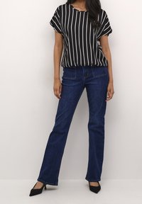 Femme portant un haut noir à manches courtes avec des rayures verticales blanches, un jean bootcut bleu foncé, et des talons noirs à bout pointu.