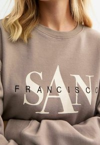 Felpa marrone chiaro con testo sovradimensionato bianco e nero che recita "SAN FRANCISCO", con una vestibilità comoda e scollo a costine.