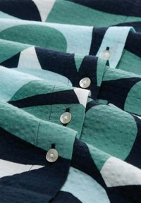 Chemise à boutons avec un motif géométrique dans des nuances de bleu sarcelle et de marine, fabriquée en tissu texturé avec des boutons en perles pour la fermeture.