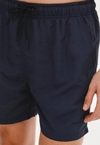 Granatowe szorty kąpielowe z elastycznym pasem, związywaniem na sznurki, kieszeniami bocznymi oraz teksturowanym wykończeniem materiału.