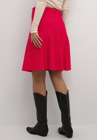 Jupe A-line rose vif, arrivant aux genoux, fabriquée en tissu lisse avec une taille plate. Portée avec des bottes en cuir noires montantes.