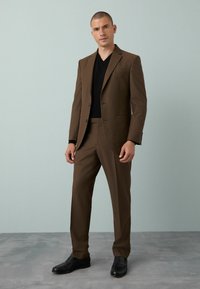 Costum maro cu un design cu un singur rând de nasturi, asortat cu o cămașă neagră cu decolteu în V. Pantofi negri completează aspectul pe un Fundal moale de gri.