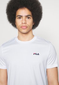 Camiseta blanca de manga corta hecha de un tejido ligero, con un pequeño logo de Fila en azul marino y rojo en el área del pecho izquierdo. Textura suave.