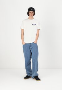 Hvit bomullst-skjorte med lilla logo, løstsittende blå denimjeans, og svart caps med logo. Sorte joggesko med brune såler.