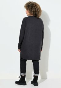 Grauer gestrickter Cardigan mit lockerer Passform, langen Ärmeln und einem Saum, der bis zur Mitte des Oberschenkels reicht, kombiniert mit dunklen Jeans und schwarzen Stiefeln.