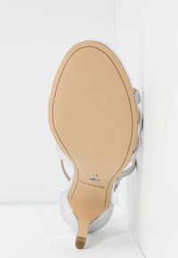 Sandale argentée à talon haut avec une tige synthétique lisse ornée d'accents pailletés, un bout pointu et une semelle en caoutchouc beige avec un talon surélevé.