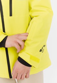 Felgeel weerbestendig jack met zwarte rits en zakdetails. Heeft een klittenband manchet en logo-accenten op de mouw.