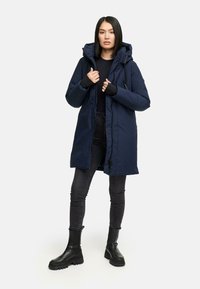 Kvinde iført en marinblå vinterjakke med hætte, sort langærmet top, mørkegrå skinny jeans og sorte klodsede støvler, der står foran en ensfarvet baggrund.