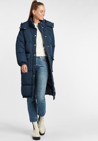 Navy blue long puffer coat with a detachable hood, large pockets, and snap closures, worn over a cream sweater and blue jeans, paired with beige boots.