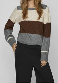 Sweter w paski w kolorze kremowym, brązowym i szarym. Ma długie rękawy, okrągły dekolt i ściągacze na mankietach. W połączeniu z czarnymi spodniami.