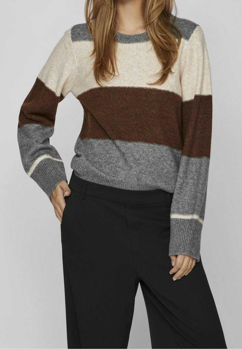 Sweter w paski w kolorze kremowym, brązowym i szarym. Ma długie rękawy, okrągły dekolt i ściągacze na mankietach. W połączeniu z czarnymi spodniami.