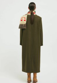 Long olive green coat with a straight cut, paired with a beige scarf featuring red scales and fringed edges, accentuating the neckline.