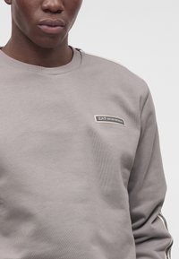 Крупен план на човек, носещ светлосиво суичър с лого EA7 Emporio Armani на гърдите и райета на ръкавите.