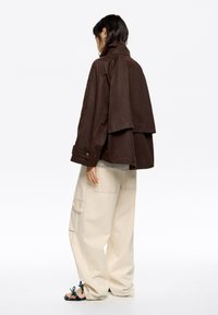 Chaqueta marrón de gran tamaño con cuello alto y diseño de espalda en capas. Combinada con pantalones de pierna ancha de color beige claro y sandalias con estampado.