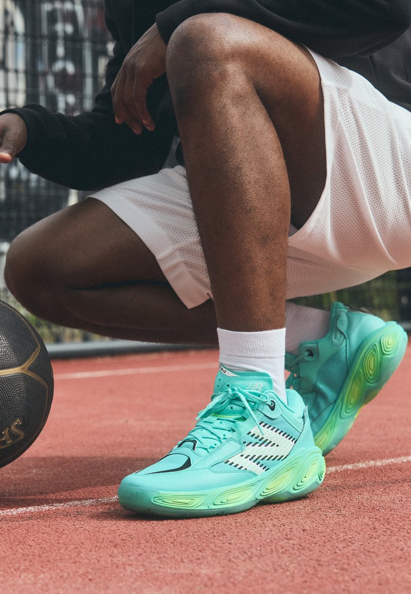 Türkise Sportschuhe mit schwarzen Akzenten und strukturiierter Sohle, mit einer glatten Oberseite und ausgeprägten Designelementen, die auf einem Platz liegen.