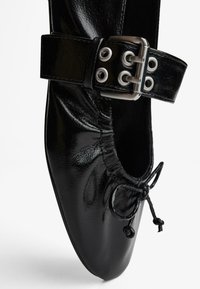 Schwarze Ballerinas mit glänzender Lackoberfläche, ausgestattet mit einem breiten Riemen mit metallenem Schnallenverschluss und Ösen sowie einer dekorativen Kordel.