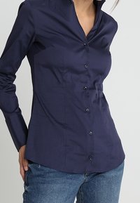 Kobieta w dopasowanej granatowej koszuli z długimi rękawami zapinanej na guziki oraz niebieskich dżinsach, na jednolitym tle.