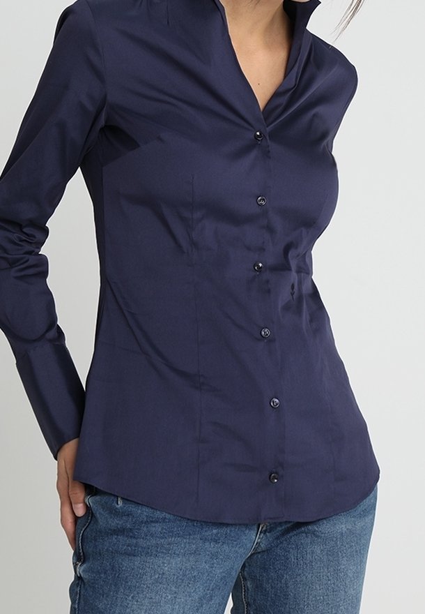 Kobieta w dopasowanej granatowej koszuli z długimi rękawami zapinanej na guziki oraz niebieskich dżinsach, na jednolitym tle.